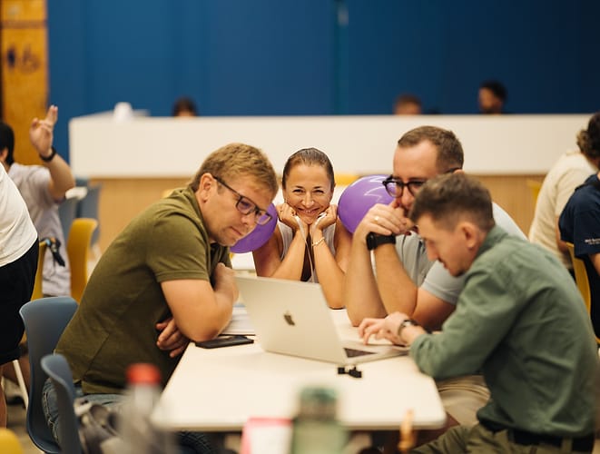 A smiling participant between two purple balloons while teammates work on a laptop